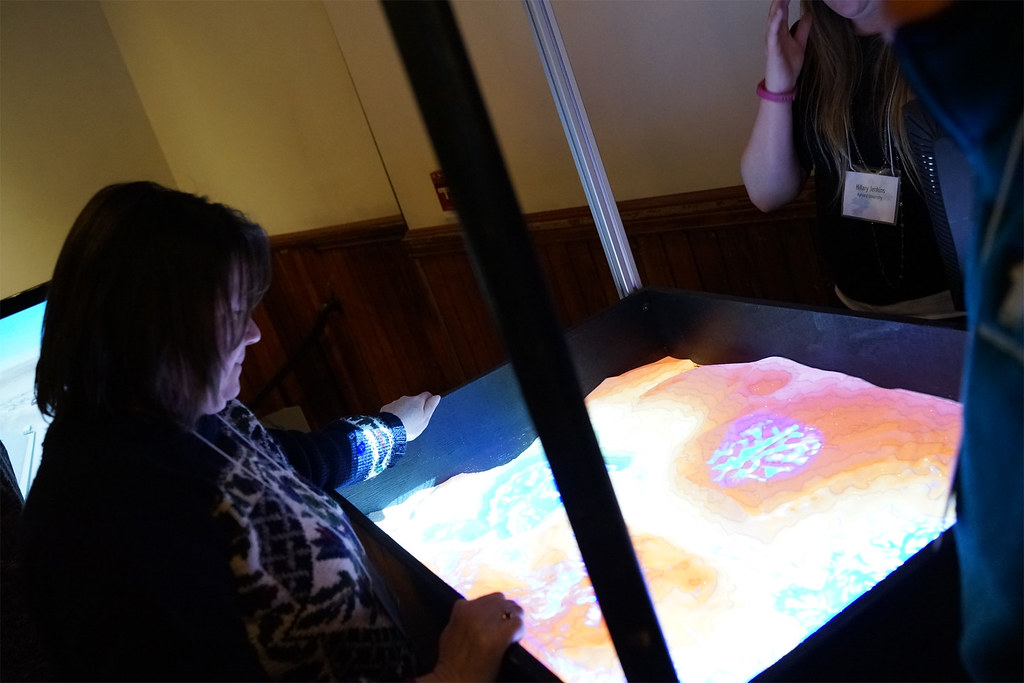 An attendee shapes real sand in an augmented-reality sandbox while a projector paints glowing elevation colors onto the terrain.