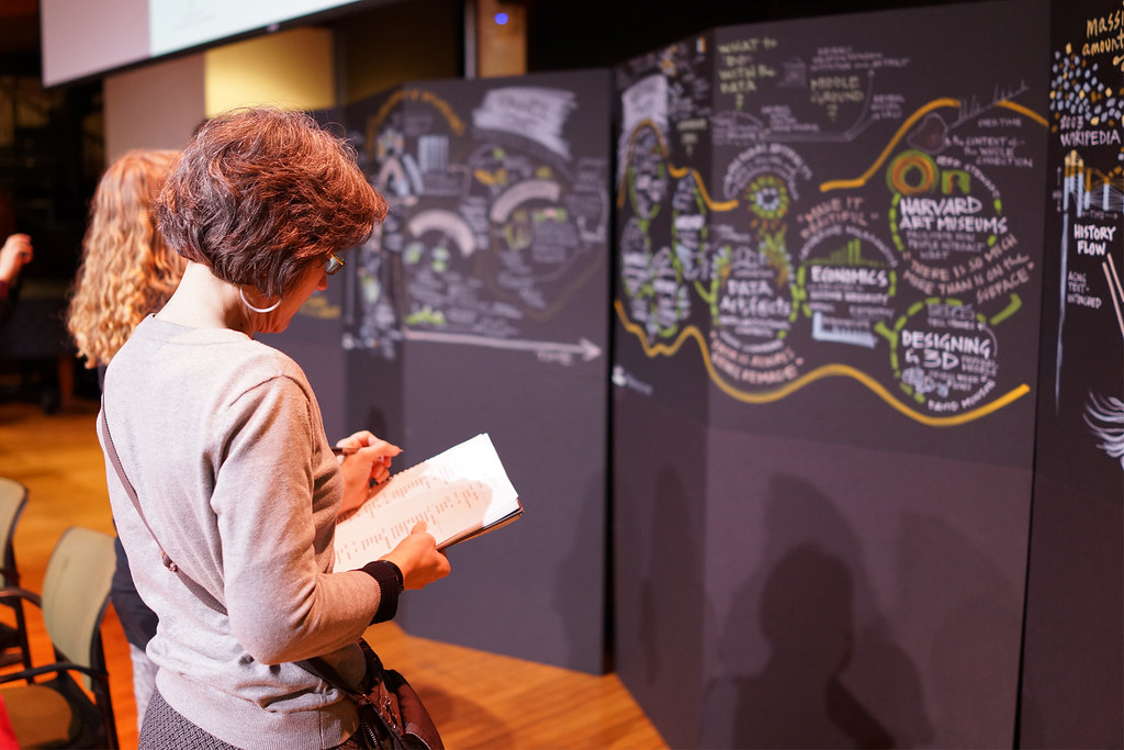 Two attendees stand close to tall black panels covered in hand-drawn graphic recordings, one taking notes on a pad.