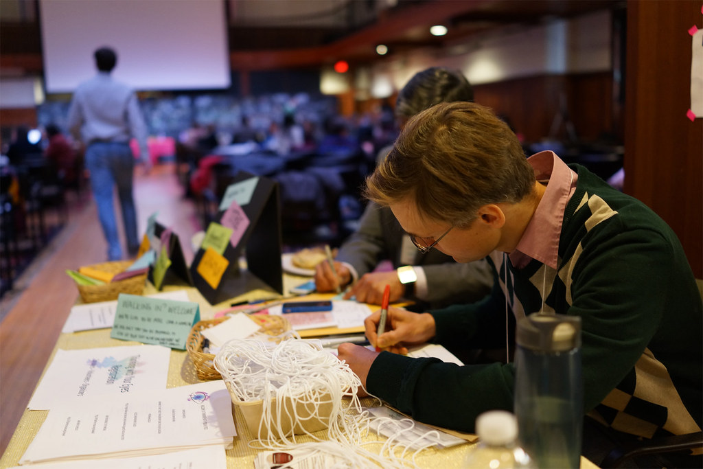 Volunteers at the greeting desk threading name-tag lanyards beside baskets of sticky notes, with the symposium hall filling behind them.
