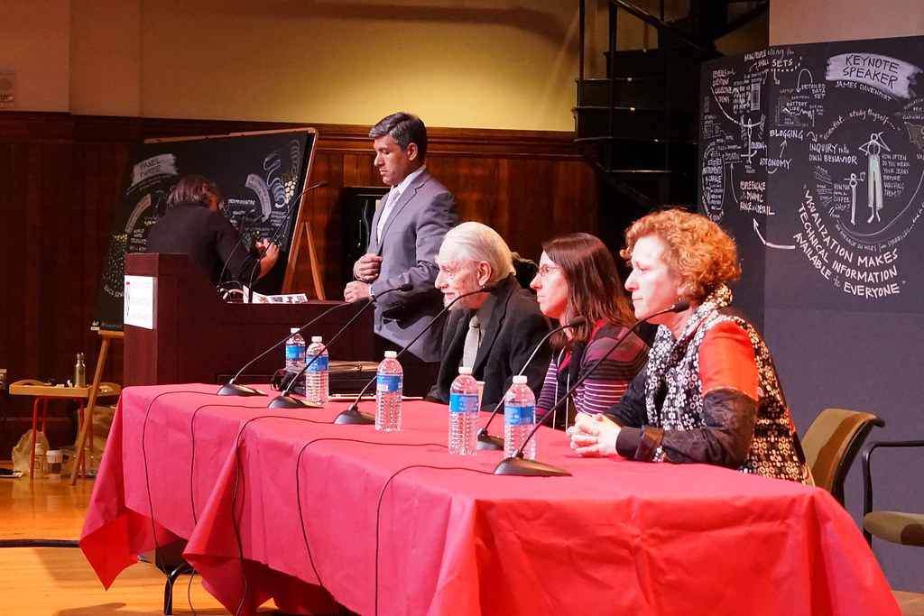 Four panelists seated at a long red-draped table on stage, framed by chalk-drawn graphic recordings of earlier sessions.